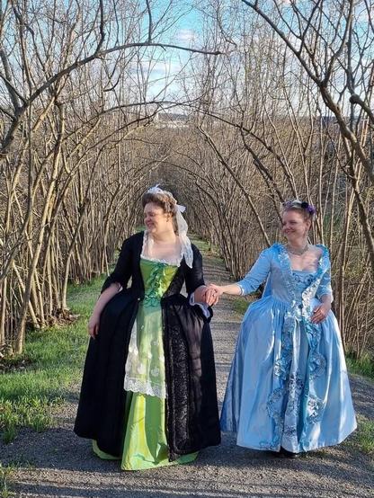 Two women wearing elaborate 18th-century-style gowns walk hand in hand down a tree-lined path. One wears a green petticoat and stomacher with a black overdress and white lace, while the other wears a pale blue gown with ruffled trim. They are smiling at each other under an archway of leafless branches on a sunny day.
