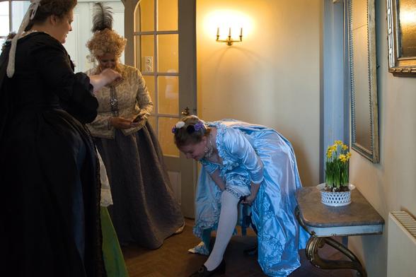 Three women dressed in 18th-century-style gowns are gathered in a warmly lit interior. One woman in a light blue dress is bending down to adjust her stocking, laughing. Another woman in a black dress stands nearby, while a third in a silver-gray outfit watches with a smile. A small table with a pot of yellow daffodils sits against the wall under a mirror.