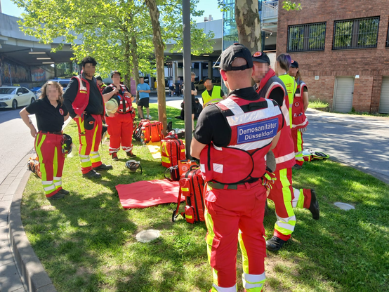 Hier zusehen ein Behandlungsplatz der Demosanitäter, einige Sanis sind auf dem Bild zu sehen, sie tragen uniform und gucken teilweise in die Kamera, auf dem Boden liegen mehrere Planen in verschiedenen Farben zur Triage sowie einige Rettungsrucksäcke und anderes Material.