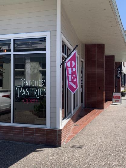 Sunny day view of a corner storefront with tall windows. One window has Patches Pastries stenciled on it. At the corner hangs a pink and white banner that says Open.