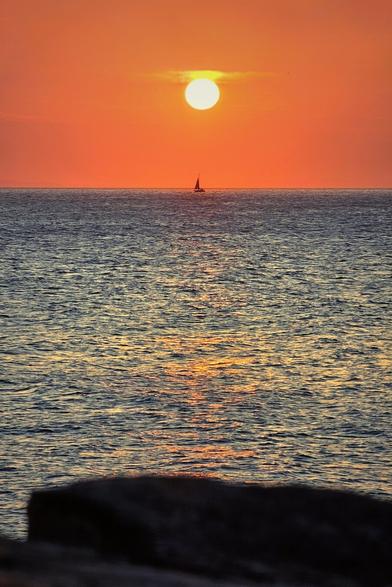 A wide shot of the ocean at sunset with a single sailboat on the horizon beneath a bright, round sun in an orange sky.
