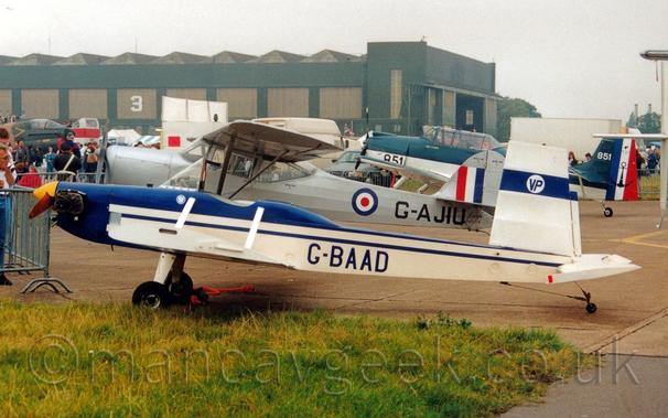 Side view of a cream and blue, long and thin single-engined light aircraft parked facing to the left.
A glazed, low windshield is the only concession to protection for the open cockpit and any pilot that may fly this.
The plane has dark blue upper surfaces and a cream body, with a white tail with a blue stripe containing a white circle with the letters "VP" in the middle in blue.
The registration "G-BAAD" are on the side of the rear fuselage, also in blue.
A pair of other single-engined light aircraft, both with military markings, are parked behind this one.
Grey metal fencing surrounds these planes, with crowds of people on the other sides, as well as a couple of vehicles.
A large green and brown hangar stretches across the frame from the left, under grim grey sky.