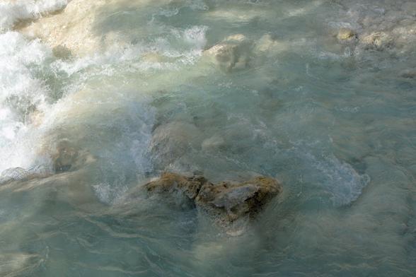 A soft, milky-blue stream flows over smooth, partially submerged rocks. Gentle currents ripple across the surface, creating a calm yet dynamic texture. Light filters through from above, casting subtle highlights and shadows on the water. [alt-text gpt-generated]