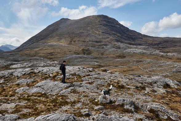 A very rocky landscape leading to a mountain peak. In the foreground Skye the border collie runs over to a guy who's probably taking some video on his gopro.