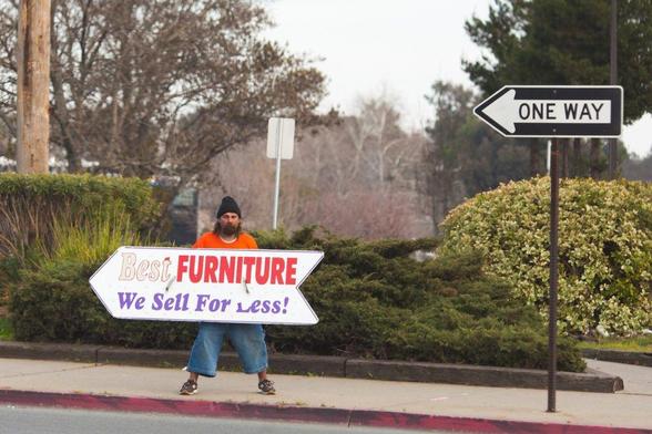 next best thing: photograph of a street with a ONE WAY sign. A man on the sidewalk is carrying a large arrow-shaped sign that says "Best FURNITURE We Sell For Less!"