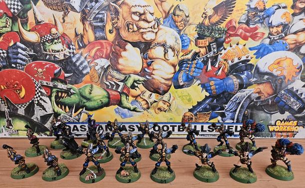 Das Foto zeigt im Vordergrund eine Aufstellung von 20 kunstvoll bemalten Miniaturfiguren eines Dunkelelfen-Teams aus dem Fantasy-Brettspiel Bloodbowl. Jede Figur steht auf einem runden, grün strukturierten Base mit einer schwarzen Zahl, die sie eindeutig identifiziert. Die Spieler tragen dunkle Rüstungen mit blauen und goldenen Akzenten sowie markanten Frisuren – viele mit langen, wilden Haaren. Einige Figuren halten Football-ähnliche Bälle, andere posieren in dynamischen Wurf- oder Fangpositionen. Im Hintergrund befindet sich die großflächige, farbenintensive und im Comicstil gehaltene Schachtel des Bloodbowl-Grundspiels: Es zeigt eine brutale Szene zwischen grünhäutigen Orks mit roten Helmen und Menschen in blauer Rüstung, die sich im Stil amerikanischen Footballs bekämpfen. Links unten ist das Citadel-Logo, rechts das bekannte „Games Workshop“-Logo zu sehen. Die Szene strahlt Energie, Humor und Fantasy-Atmosphäre aus.