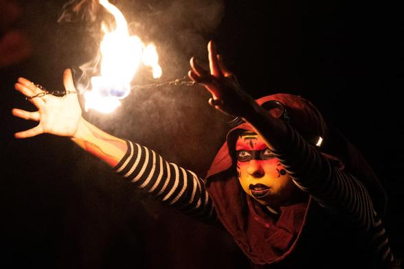 A person with a painted face twirls a fiery ball between their hands.