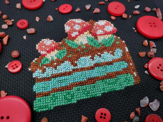 A cross stitch of a green ombre cake slice with brown frosting, topped with red and white mushrooms and leafy greens on black aida. The cross stitch is surrounded by red buttons and small, red translucent rocks.