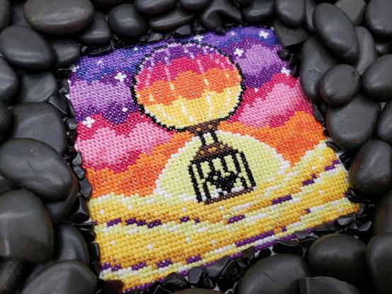 A cross stitch of a silhouetted bird in a cage floating along against a backdrop of a colourful sunset  of pinks, purples, and oranges. The cross stitch is surrounded on all sides by shiny black stones.