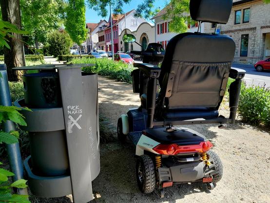 The scoot-chair is parked next to a trash can emblazoned with FUCK NAZIS, and the Shitter logo, which is a sans-serif swastika.