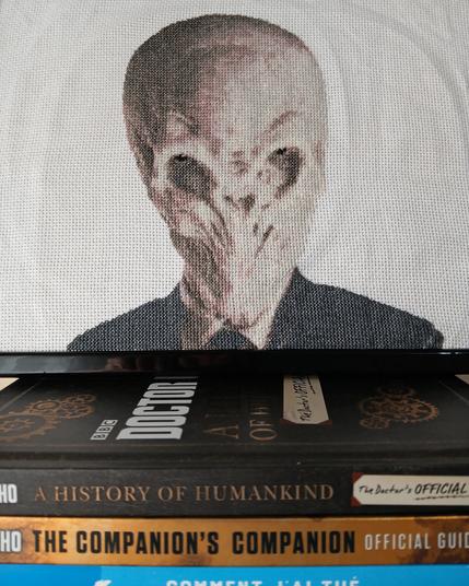 Photo d'un point de croix dont le thème est la série Doctor Who.
Le cadre est posé sur des livres anglais parlant de la série, ainsi que sur un livre bleu dont on voit un bout de tranche. (Il s'agit de "Comment j'ai tué mon ange gardien" de Thomas Hercouët. Et c'est une private private joke, dans le sens où je me suis fais la blague que l'ange du livre pourrait être un nouveau Docteur mais cette blague n'est jamais sortie de ma tête, jusqu'à présent! Bref!)

Il représente un extraterrestre humanoïde dans un costard (Oui alors je fais ce que je peux pour la description mais avec Doctor Who il vaut mieux éviter de se poser trop de questions). Sa tête est allongée avec le crâne assez gros. Ses orbites sont très creuses et larges mais il a des yeux de tailles humaines.
Il n'a pas de nez, plutôt une cavité, et pas de bouche non plus.
Il est donc habillé avec ce qui ressemble à un costard, avec petite chemise blanche dont le col dépasse, parce que la série est anglaise donc l'élégance est de mise, vous comprenez! 

Le point de croix ne représente que la tête et le haut des épaules. Le fond est blanc.