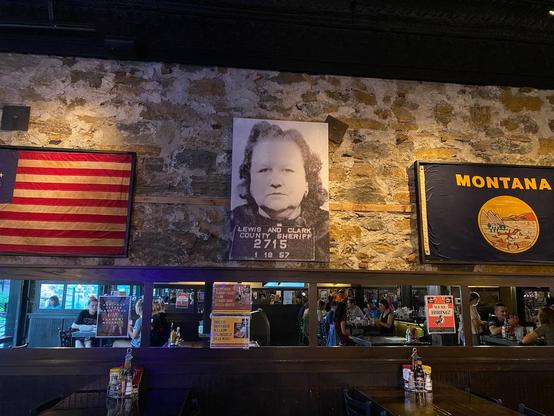 Photograph of a wall behind a restaurant bar. On the wall are framed flags of the United States and the sate of Montana (both historical flags). In between the flags is a mug shot of Big Dorothy.