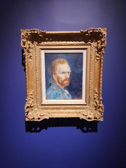 In an elaborate gold frame, an 1887 self portrait of Vincent van Gogh. He wears a blue jacket and is looking to his right at us. His hair is short, beard of medium length, both red in color.