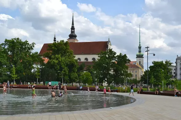The image shows a park area with a fountain or reflective water feature where people are enjoying themselves, some wading or playing in the water. In the background, there are trees and buildings with notable architectural features, including church spires. The sky is partly cloudy, suggesting a pleasant day for outdoor activities.

Image Credits: Wikimedia / RomanM82 / CC BY-SA 4.0