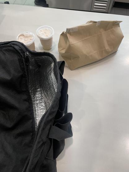 A black food delivery bag sits partially open on a store counter. Beside it is a small brown food bag and two small brown frosty drinks.