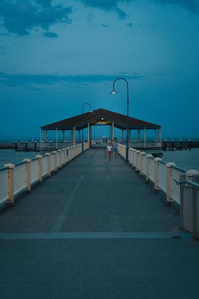Photo of a jetty at dusk in Redcliffe, Queensland, Australia. The sky is a little hazy and two people can be seen walking towards the camera in the distance