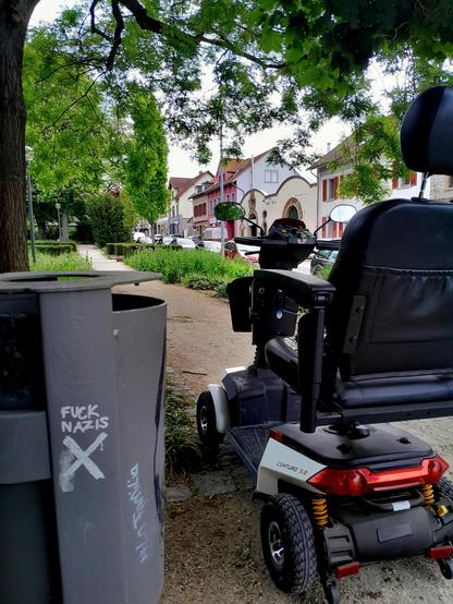 The scoot-chair is parked next to a trash can emblazoned with FUCK NAZIS, and the Shitter logo, which is a sans-serif swastika.
