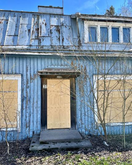 Abandoned blue house with peeling paint, boarded windows, and a wooden door near the entrance, surrounded by overgrown bushes.