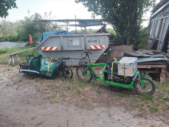Ein grünes Bullitt Lastenrad mit einigen Kisten auf der Ladefläche und einem Carla Cargo Anhänger steht vor einem Container voller Eselmist. Auf dem Anhänger sind zwei mit Mist gefüllte Laubsäcke.