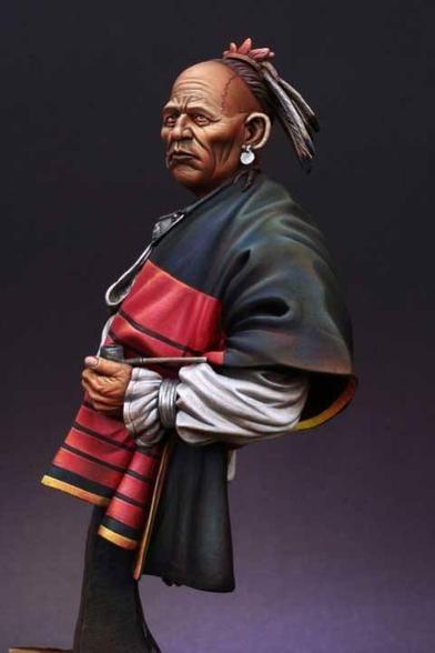 This image presents a masterfully crafted and painted miniature sculpture of a Native American elder, striking a graceful yet commanding pose. The composition exudes a sense of cultural pride, strength, and dignity, with the figure's expression and posture inviting contemplation of their life and heritage. The figure's accessories further enhance its storytelling. Large silver earrings glint softly, and the feathers in his headpiece rise gracefully, adding both height and symbolic weight to the sculpture. The feathers' delicate details suggest a connection to nature or spirituality, while the pinkish ornament at the crown adds an unexpected splash of color that enlivens the overall palette. His hands hold the shawl firmly yet gently, creating a subtle dynamic that conveys both a sense of action and stillness.