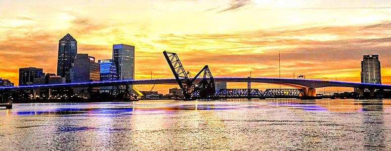 Originally built in 1921, and replaced in 1991. 1,645 feet, connecting downtown Jacksonville with the Southbank of the Saint Johns River. Known for the neon strip lighting down the bridge center, originally blue, now a variety of colors for different occasions.  Here seen during a colorful morning sunrise with the downtown skyline in silhouette against bright shades of yellow and orange filling the sky beyond the bridge, with the blue neon strip adding to the colorful reflections upon the water below.