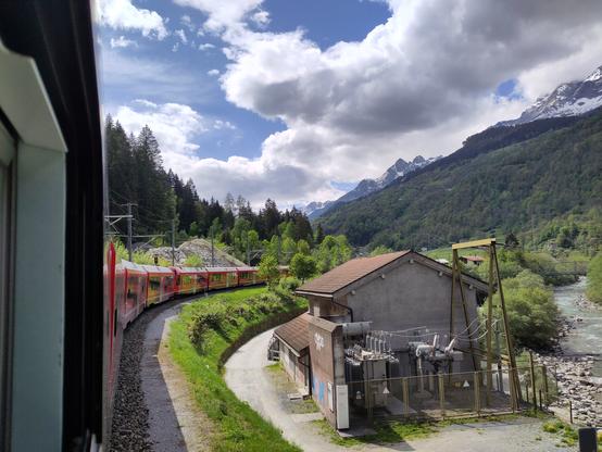 Foto. Ein roter Zug in Berglandschaft mit einem Kraftwerk.