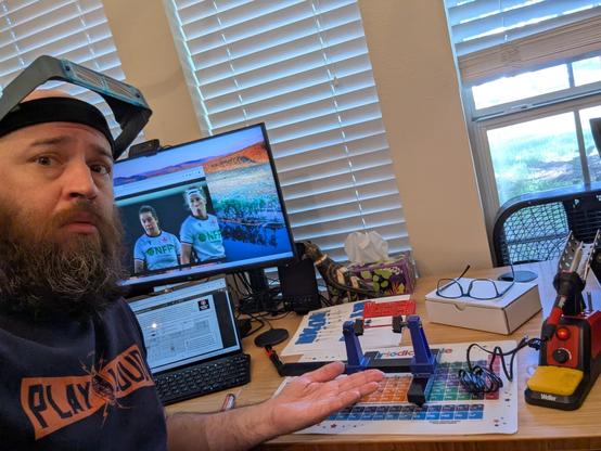an adult white man with a beard wearing a blue T-shirt that says "Play Loud" and a magnifying visor with a concerned facial expression gestures at the desk before him. on the desk is a soldering station, a PCB holder with a small red PCB on it, a sheet with electrical components taped to it, the manual for the kit on one computer monitor, and USA v Canada Women's rugby on the other monitor.