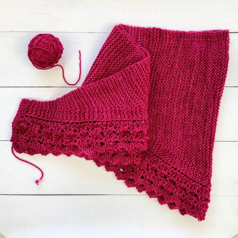 Flat lay view of the remaining ball of dark pink yarn, and a knitting project featuring garter stitch and lace. It's shown on a white plank background.