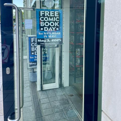 A glass storefront door displaying a sign for "Free Comic Book Day," indicating it will occur on May 3, 2025, the first Saturday in May.