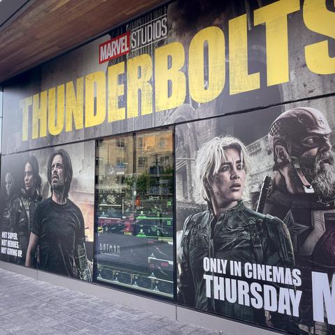 A promotional poster across a comic book shop window for "Thunderbolts" by Marvel Studios. The design features the title "THUNDERBOLTS" prominently in yellow, alongside images of several characters, including a woman with short blonde hair and a man in a black shirt.