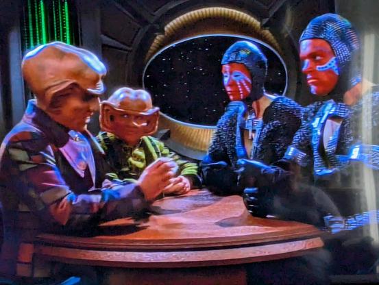 Aliens chatting around a table. Two of them look like oompa loompa on steroids