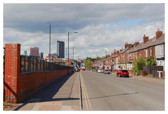 This image depicts a quiet urban street lined with red-brick terraced houses on the right side. Each house has a small front yard, and there are parked cars along the street. On the left, a tall brick wall runs parallel to the sidewalk, with a metal fence on top. In the distance, the street gently slopes downward, leading toward a city skyline with several tall buildings, including modern skyscrapers. The sky is partly cloudy, with patches of blue visible. The scene is calm, with a few pedestrians walking along the sidewalk.