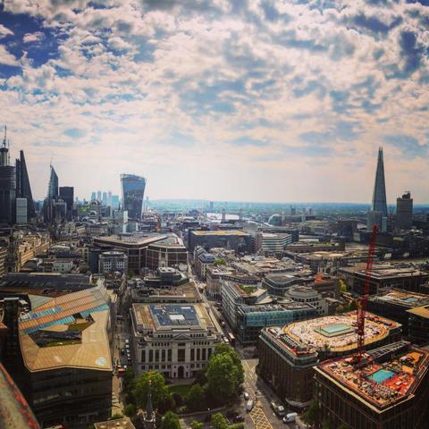 The image shows a panoramic view of London's cityscape, highlighting a number of iconic skyscrapers and architectural landmarks. Prominent in the picture are the 20 Fenchurch Street (commonly known as the "Walkie-Talkie"), the Leadenhall Building (often referred to as the "Cheese Grater"), and The Shard, known for its distinct spire-like shape. The skyline shows a mix of modern glass-covered office buildings and older historic structures, set against a backdrop of a partly cloudy sky. Additionally, the River Thames is visible in the distance, alongside other familiar elements of London's vast urban landscape.
