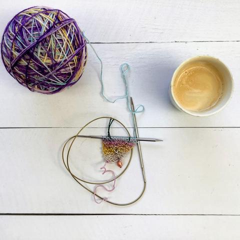 Flat lay view of a ball of variegated yarn, a circular knitting needle with the start of a knitting project, and a cup of coffee on a white plank background.
