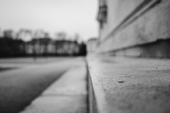 Monochrome photo with shallow depth of field showing the textured edge of a stone ledge or pathway in sharp focus, while the background with bare trees and a building fades into a soft blur.
