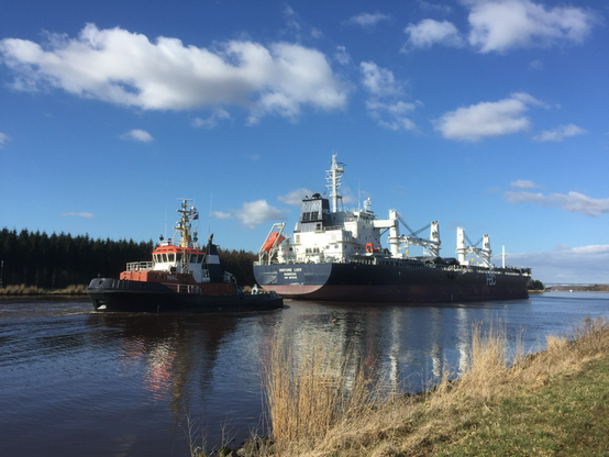 Farbfotografie im Querformat zeigt ein großes Containerschiff (4 Krane), das auf dem Nord-Ostseekanal unterwegs ist. Am Heck ist ein Schlepper vertäut (in entgegengesetzter Fahrtrichtung). Der Himmel ist blau mit wenigen Wölkchen.