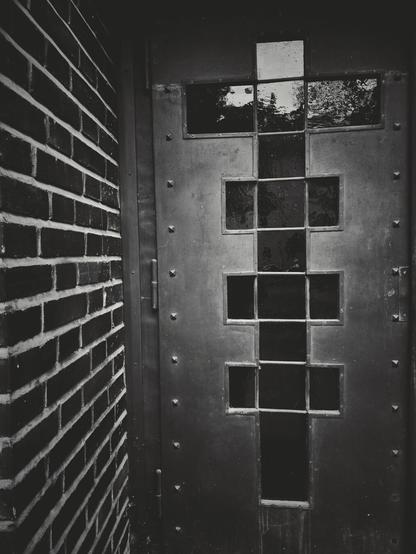 The portrait-orientated black and white image shows a close-up view of a brick wall and a metal door. The wall is on the left side of the image and is constructed of rectangular bricks laid in a pattern. The metal door is on the right side of the image. It is made of solid material with visible rivets or fasteners. The door has a pattern of multiple small, rectangular glass panes. The panes are arranged in a grid-like structure that forms a cross shape.