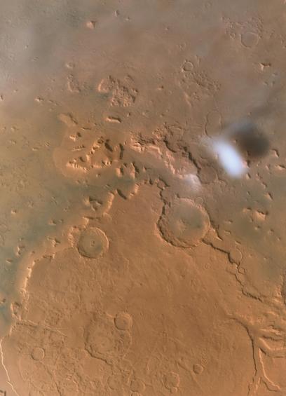An orbital image of the Martian surface taken by Tianwen-1, showing the region of Protonilus Mensae in the Ismenius Lacus quadrangle. The landscape features a mix of rugged, eroded mesas and numerous impact craters. Near the bottom, an eroded landform resembles the shape of an ancient riverbed. The terrain varies in colour from warm rusty reddish tones to more brownish, with contrasting light and dark shading that reveals surface textures and elevation changes. Very light cloud cover is visible near the top of the image, softening the details in that region. Toward the right side, a distinct, bright white cloud casts a soft shadow, standing out sharply against the rusty ground. The overall scene conveys the dry and rocky, yet ancient and geologically complex nature of the Martian terrain.