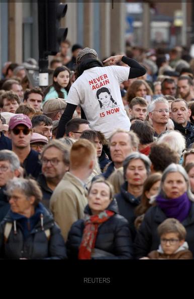 Een groep witte mensen kijkt voor zich uit. Er komt iemand omgedraaid bovenuit met een wit shirt aan met daarop in letters NEVER AGAIN IS NOW en een foto van Hind Rajab.