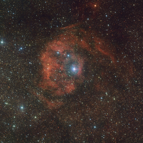 The image shows a giant red nebula in space. Behind the nebula there are countless stars of different colours and sizes. Most stars are small and have a white to pale blue hue. Overall, the nebula is round and appears to change from diffuse to concentrated towards the centre of the picture. Its colour also changes from a dark red to a vibrant orange. Within the centre of the picture, inside the brightest region of the nebula, lies a large, white to pale blue star.