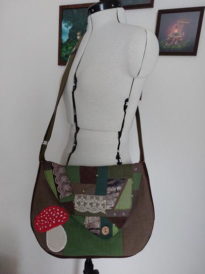 Photo d'un mannequin de couture réglable blanc, devant un mur blanc où sont accrochés des tableaux à l'ambiance forestière réalisés par Lindenshield Arts qui est sur mastodon ! 

Le mannequin porte un sac en bandoulière dans les tons verts et bruns. Il est réalisé à partir de chutes de lin et de galons en dentelle. 
Il se ferme grâce à un bouton en bois, et est orné sur le devant de morceaux de lin assemblés pour représenter une amanite tue mouche.