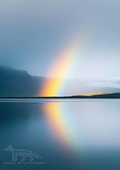 A radiant rainbow arcs from a misty mountain ridge into the still waters of a remote Icelandic fjord. The vivid colors of the rainbow reflect perfectly on the mirror-like surface of the lake, doubling the visual impact. The distant mountains are shrouded in fog, while soft light touches a grassy ridge on the right, enhancing the ethereal, dreamlike quality of the scene.