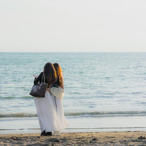 The image shows two people standing on a beach facing the ocean. They appear to be close to each other, possibly embracing or posing. One of them has a tote bag. The setting is serene, with calm waves and a clear sky.