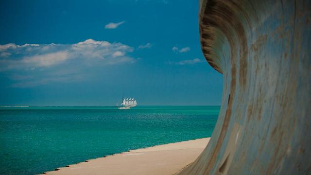 Photo of a multi-masted sailboat and a sailboat with sails down in the blue/green waters of Lake Michigan. Clouds are seen in the distance. In the foreground a seawall, curved at the top and rusty throughout is seen next to a concrete sidewalk/bike trail. The seawall looks similar to a wave rushing towards the distant sailboat.