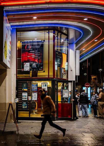 Close-up shot on the entrance of Art Deco style Cine vox cinema in Strasbourg at night, illumitated by its colourful red, blue and yellow neons. A young man is running at the front of the building.