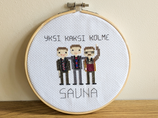 A small cross stitch project on white fabric, framed in a wooden hoop and sitting on a wooden shelf. It features three men in suits and ties, one holding a mic and grinning widely, another with glasses, a moustache and a red accordion, and the third guy in the middle has his arms around the shoulders of the other two. The text above and below them reads "Yksi kaksi kolme sauna", Finnish for "one two three sauna".