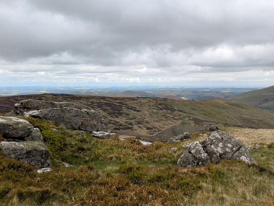 Colour photograph of a rocky outcrop, the summit of the Schil. Views over distant valleys and hills. Grey cloudy sky.