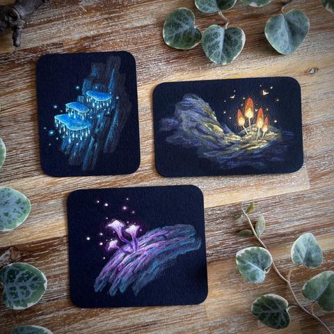 A photo of three small paintings on black paper with rounded corners, placed on a wooden table and surrounded by leaves and branches for decoration. The top left shows shelf-like teal mushrooms with tiny glowing beads hanging off the rims; the top right paintings shows simply cone-shaped orange glowing mushrooms illuminating a rocky surface, a few tiny insects hovering around them; the bottom painting shows pink, trumpet-shaped mushrooms, a sphere-like structure on their top glowing softly.
