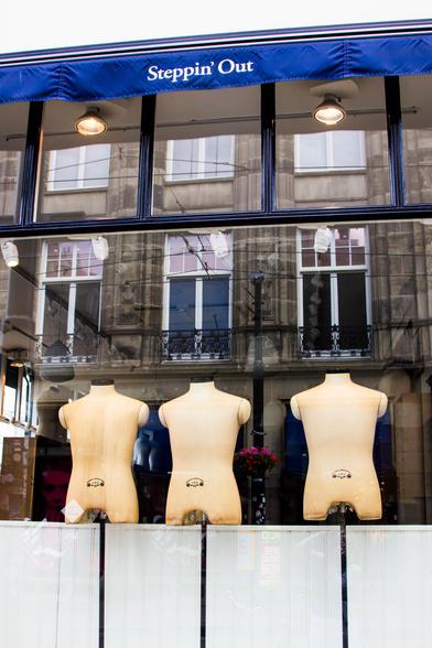 A shop window with three mannequins in it, torsos only and naked. They are just above a horizontal white screen of 50 cm. approximately. Exactly above the torsos the reflection of the windows of a historic building across the street, exactly above each torso one. At the top a strip of a blue sunshade with white letters: "Steppin' Out".
