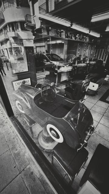 The image is a black and white photograph depicting a view through a window with a reflection. The reflection on the window shows a street scene with buildings, a sidewalk, and a pedestrian. Visible through the window is the interior of a barbershop, containing chairs and various items. A small, old-fashioned car model of a barber chair for kids is placed inside the window display. Several diplomas, one of which reads "DIPLOM," are visible on the window's side wall.